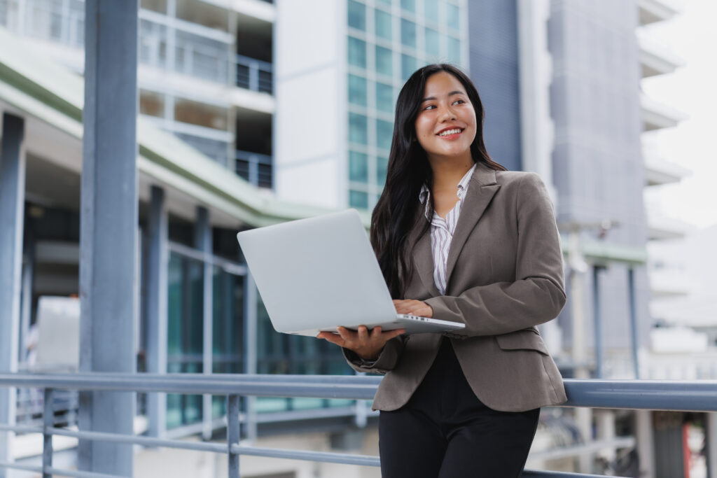 A young corporate professional holding her laptop and standing confidently outside her office.