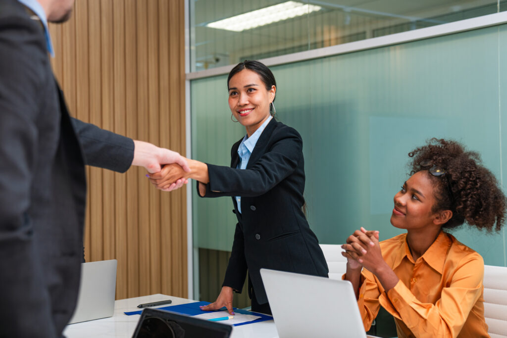 Two business professionals shaking hands with another professional sitting down and looking up at them.