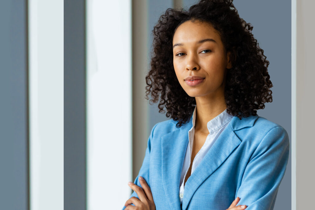 A young professional in business attire standing in a modern office setting with her arms crossed